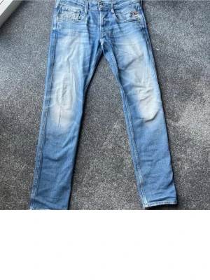 Ljusblå jeans från Replay - Säljer ett par ljusblå jeans från Replay med snygga slitningar och klassisk femficksmodell. Jeansen har raka ben och detaljer i form av Replay-logga vid fickan. Perfekta för en avslappnad och trendig stil.