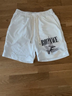 Vita “ROMANCE” shorts med duvmotiv🤍 -  Snygga och bekväma vita shorts med elastisk midja och dragsko för perfekt passform. Stilren design med trycket “ROMANCE” och detaljerat duvmotiv som ger en trendig och unik look. Mjukt och lätt material som passar perfekt till både vardag, semester och avslappnade outfits.  * Färg: Vit * Elastisk midja med dragsko * Trendigt print framtill * Bekväm passform * Perfekta för sommar och streetwear-stil