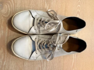 Vita sneakers från Lorenzo - Stilrena vita sneakers från Lorenzo med klassisk låg profil och vita snören. Skorna har en enkel design med diskret logga på hälen och ovandel i slätt läder. Perfekta för en clean och avslappnad look.