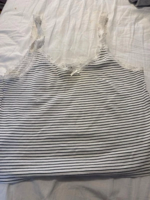 Randig vit linnetopp med spets - Söt vit linnetopp med tunna svarta ränder, spetsdetaljer längs urringningen och axelbanden samt en liten rosett framtill. Perfekt för en avslappnad och trendig look.