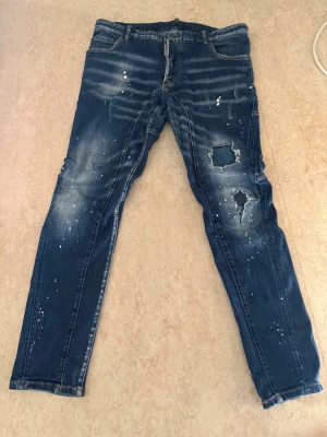Dsquared2 blå slitna jeans herr - Säljer ett par blå jeans från Dsquared2 med coola slitningar, hål och färgstänk. Jeansen har smal passform, klassisk femficksdesign och tydliga detaljer på benen. 