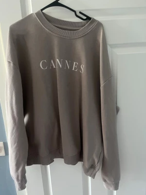Beige sweatshirt med tryck 'CANNES' - Snygg beige sweatshirt med texten 'CANNES' tryckt framtill. Tröjan har rund halsringning, ribbade muddar vid ärmslut och nederkant samt en avslappnad passform. Perfekt för en chill och stilren look.