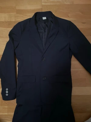 Mörkblå kostym med kavaj och byxor - Snygg mörkblå kostym med klassisk kavaj och matchande byxor. Kavajen har två knappar framtill och dekorativa knappar vid ärmslutet. Har använt två gånger😄 Storlek fins i bilder, priset kan diskuteras 