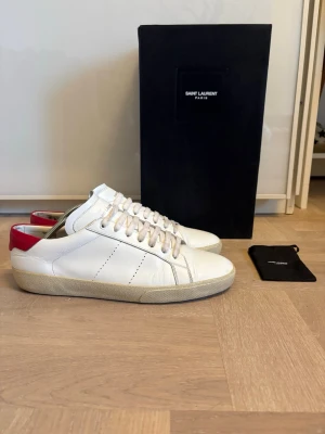 Saint laurent skor - Snygga vita sneakers från Saint Laurent i läder med röd detalj på hälen och beige sula. Klassisk låg modell med vita skosnören och diskreta sömmar. Perfekta för dig som gillar stilrena och trendiga skor. Box ingår