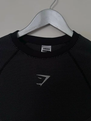 Gymshark onyx v5 - Säljer en svart långärmad sporttröja från Gymshark med diskret mönster och logga på bröstet. Tröjan har rund hals och är tillverkad i ett tekniskt material som andas, perfekt för träning och aktivitet.