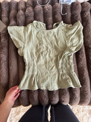 Ljusgrön blus med smockdetaljer - Säljer en ljusgrön blus med smockdetaljer i midjan och korta, vida ärmar. Blusen är luftig och skön nu i sommar. 