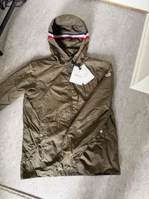Olivgrön Moncler vindjacka med huva - Snygg olivgrön vindjacka från Moncler med huva och dragkedja. Jackan har Moncler-logga på ärmen och detaljer i rött, vitt och blått på huvan. Materialet är lätt och vattenavvisande, perfekt för att hålla sig torr. Två fickor med knappstängning och justerbar huva.