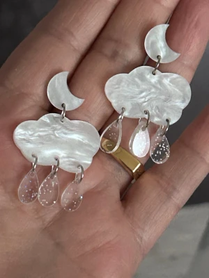 Molnformade örhängen med måne - Unika örhängen i form av vita moln med en halvmåne upptill och hängande transparenta droppar som liknar regn. Materialet är skimrande och ger ett coolt, drömmigt intryck. Perfekt för dig som gillar statement-smycken och fantasifulla accessoarer.