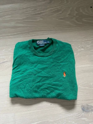 Grön stickad tröja Polo Ralph Lauren - Snygg grön stickad tröja från Polo Ralph Lauren i slim fit-modell. Tröjan har rund hals och klassisk orange logga broderad på bröstet. Perfekt för dig som gillar stilrena och tidlösa plagg. Materialet är merinoull.