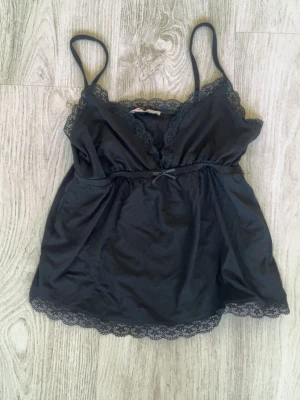 Svart spetslinne  - Snyggt svart linne från Nelly i satinliknande material med tunna axelband och spetsdetaljer vid urringning och nederkant. Söt liten rosett framtill och figurnära passform🖤Kolla in min profil för fler fina och trendiga plagg!