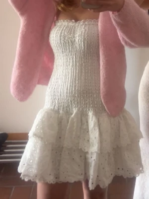 Vit kort klänning med volanger - Säljer en söt vit kort klänning med smock upptill och två lager volanger nertill. Klänningen är ärmlös och har broderade detaljer i tyget som ger en romantisk känsla. Perfekt för sommarens alla tillfällen!