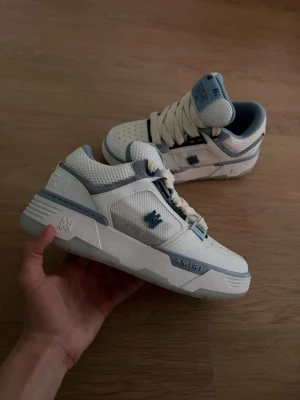 AMIRI vita och blå sneakers - Snygga AMIRI sneakers i vitt och ljusblått med mesh och läderdetaljer. Skorna har stjärnmönster på tån, breda vita snören och tydliga AMIRI-loggor på sidan och plösen. Låg modell med chunky sula och coola detaljer som ger en modern vibe.
