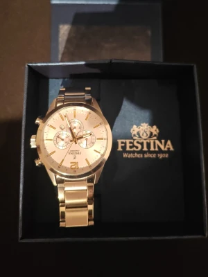 Silverfärgad kronografklocka Festina - Stilren kronografklocka från Festina i silverfärgat rostfritt stål. Urtavlan är vit med guldfärgade detaljer och tre mindre subdials. Länken är klassiskt polerad och klockan har tydliga knappar på sidan för funktioner. Perfekt accessoar för en modern look.