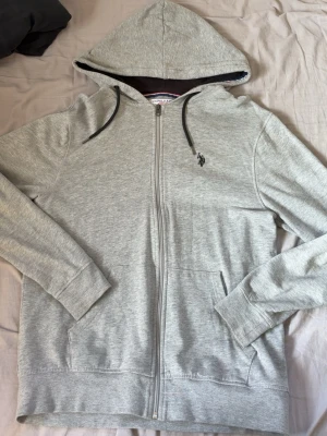 Grå hoodie från U.S. Polo Assn - Säljer en grå zip-hoodie från U.S. Polo Assn med svart dragkedja och broderad logga på bröstet. Hoodien har två fickor fram och är tillverkad i mjukt bomullsmaterial. Väldigt fint skick, använder ej PGA att den är för stor. Pris kan diskuteras 🤝