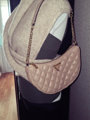 Beige quiltad handväska från Guess - Säljer en beige quiltad handväska från Guess med guldfärgade detaljer och kedja. Väskan har en halvmåneform och ett metalltriangelmärke på framsidan. Perfekt för dig som gillar trendiga accessoarer och stilren design.