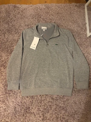 Lacoste half-zip - Snygg grå tröja från Lacoste med halvzip och klassisk krokodillogo på bröstet. Regular fit och ribbade muddar vid ärmslut och nederkant. Perfekt för en chill och stilren look, passar till jeans eller joggers.