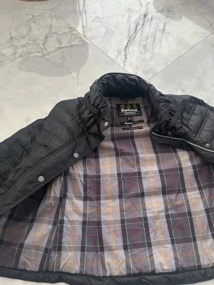  pufferjacka från Barbour -  pufferjacka från Barbour International med klassiskt rutigt foder i grått, gult och rött. Jackan har hög krage, dragkedja och tryckknappar. Perfekt för kalla vinterdagar och ger en stilren look.