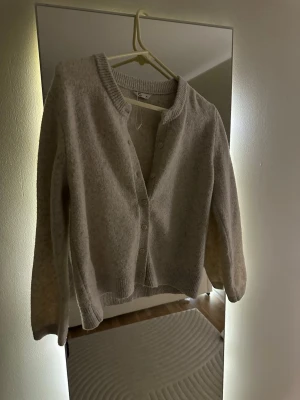 Beige kofta med knappar - Mysig beige kofta med rund hals och knäppning framtill. Koftan har långa ärmar och ribbade muddar vid ärmslut och nederkant. Perfekt att slänga på sig när det är lite kyligt. Materialet ser ut att vara mjukt och stickat.