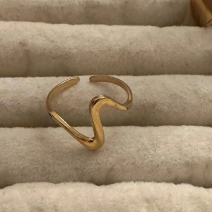 Guldigt armband med vågig design - Säljer ett snyggt armband i guldfärgad med unik vågig form. Armbandet är öppet och har en modern, minimalistisk look som sticker ut. Perfekt för dig som gillar trendiga accessoarer med coola detaljer.