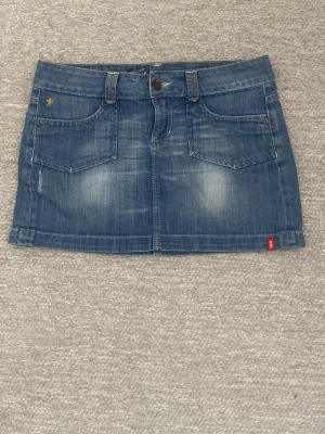 Blå jeanskjol från Replay - Snygg blå jeanskjol från Replay med klassisk femficksdesign, bälteshällor och knappstängning. Kjolen har dekorativa bakfickor med broderi och en liten Replay-logga framtill. Perfekt för en avslappnad och trendig look.