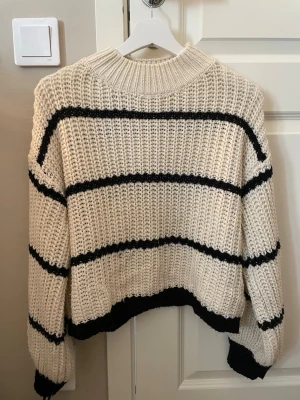 Vit stickad tröja med svarta ränder - Säljer en stickad tröja i offwhite med breda svarta ränder. Tröjan har en hög ribbad krage och ribbade muddar vid ärmslut och nederkant. Perfekt för att skapa en avslappnad och trendig look. Materialet är tjockt och mjukt.