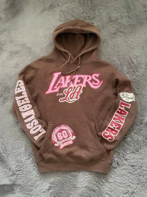 Mitchell & ness Lakers - Cool brun hoodie med stor rosa och vit Lakers-logga på bröstet och LA-tryck. Texten 'LOS ANGELES' och 'LAKERS' finns på ärmarna, samt patch med '60th Finals'. Klassisk huva och magficka. Perfekt för basketfans som vill sticka ut.