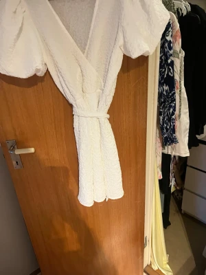 Vit omlottklänning med puffärm - Säljer en kort vit omlottklänning med puffärmar och knytband i midjan. Klänningen har en v-ringad urringning och är tillverkad i ett strukturerat tyg som ger en lyxig känsla. Perfekt för sommarens alla tillfällen!