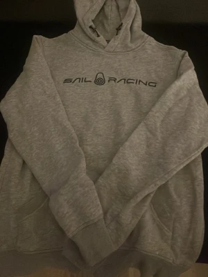 Grå hoodie från Sail Racing - Säljer en grå hoodie från Sail Racing med svart logotyp tryckt på bröstet. Klassisk modell med huva och känguruficka framtill. Mjuk och skön bomullsblandning, perfekt för chill dagar eller när du vill ha en sportig look.