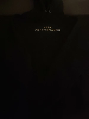 Svart hoodie från Peak Performance - Snygg svart hoodie från Peak Performance med vit logga tryckt på bröstet. Klassisk modell med huva och dragsko, samt mjukt och varmt material på insidan. Perfekt för en avslappnad och sportig stil.
