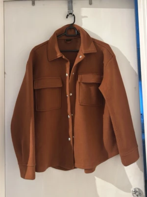 Brun overshirt med fickor - Snygg brun overshirt med klassisk krage och tryckknappar framtill. Två stora bröstfickor med lock ger en trendig look. Overshirten har raka ärmar och är tillverkad i ett mjukt, tjockare tyg som passar perfekt till lager-på-lager-stil.