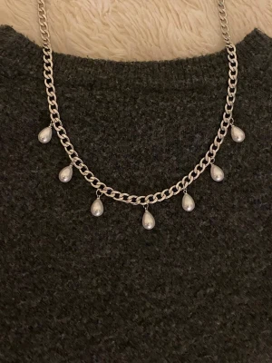 Silverfärgat halsband med pärldroppar - Säljer ett silverfärgat rostfritt halsband med kedja och sju droppformade pärlor som hänger längs kedjan. Halsbandet har en chunky kedja och pärlorna är vita och glänsande. Perfekt för dig som gillar stilrena och trendiga accessoarer.