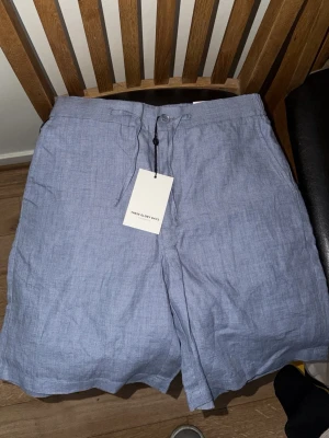 Blå linneshorts från These Glory Days - Säljer ett par ljusblå linneshorts från These Glory Days med snörning i midjan och klassisk knappstängning. Perfekt för dig som vill ha en luftig och stilren look under varma dagar. Shortsens material är linne och de har en relaxed fit.