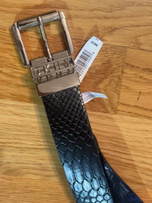 Svart bälte från DKNY med ormmönster - Snyggt svart bälte från DKNY med silverfärgat spänne och präglat ormmönster. Bältet har en bred design och logotypen är ingraverad på metallspännet. Perfekt för att lyfta vilken outfit som helst. Ny skick, äkta.
