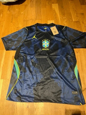 Blå Brasilien fotbollströja Nike - Snygg blå och svart fotbollströja från Nike med Brasilien CBF-logga och gula detaljer. Tröjan har korta ärmar, rund hals och inslag av grönt och gult på sidorna. Tillverkad i lätt och ventilerande material, perfekt för träning eller match.