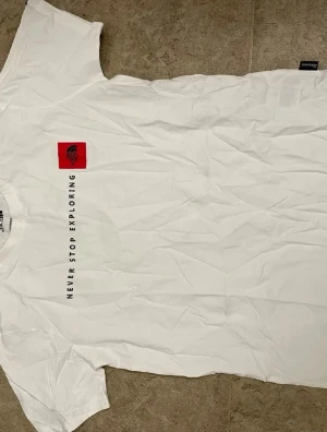 Vit t-shirt från The North Face - Vit t-shirt från The North Face med svart text 'Never Stop Exploring' och en röd logopatch på bröstet. Klassisk passform och korta ärmar, perfekt för dig som gillar enkel och stilren design. Materialet är mjuk bomull.