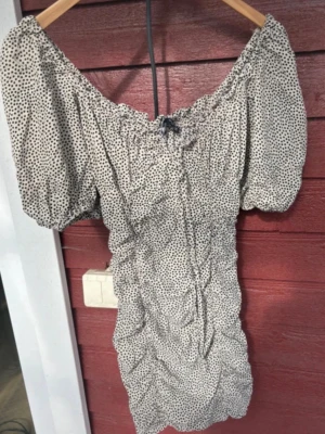 Prickig offshoulder kortklänning XS - Supersöt kortklänning med offshoulder och puffärmar i beige med svarta prickar. Klänningen har smockad midja och rynkade detaljer framtill. Perfekt för sommarens alla tillfällen och ger en romantisk vibe.