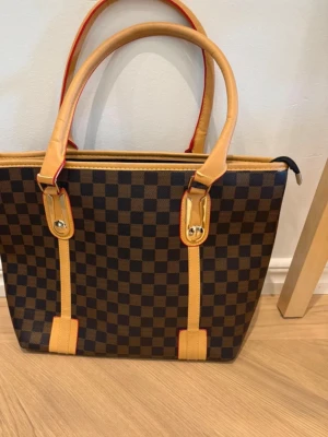 Brun rutmönstrad handväska - Snygg handväska med klassiskt brunt och mörkbrunt rutmönster, beige handtag och detaljer samt röda kanter. Väskan har två långa handtag och medföljer en liten matchande clutch. Perfekt för dig som gillar stilrena accessoarer.