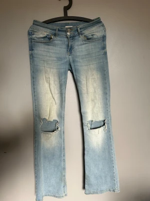 Ljusblå bootcut jeans med slitna knän - Ljusblå jeans med bootcut passform och slitna detaljer på knäna. Klassisk femficksdesign och knappstängning framtill. Jeansen har en blekt look och är tillverkade i denim. Perfekt för dig som gillar en avslappnad och trendig stil.