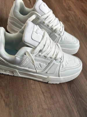 Vita Louis Vuitton sneakers - Snygga helvita Louis Vuitton sneakers med präglade monogramdetaljer och kraftig sula. Skorna har lågt skaft, tjocka vita snören och är tillverkade i en mix av läder och textil. Perfekta för dig som vill ha en clean och exklusiv look.
