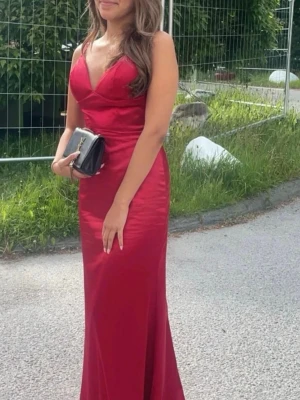 Röd långklänning i satin - Elegant röd långklänning i glansigt satinmaterial med smala axelband, v-ringning och dekorativa knappar i ryggen. Figurnära passform som framhäver siluetten och öppen rygg för en snygg look.