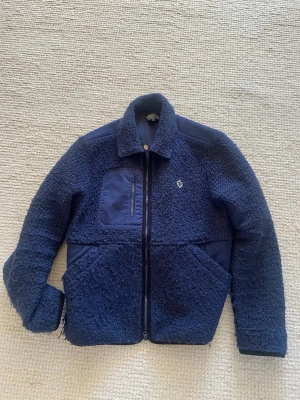 Marinblå teddyjacka från Morris - Säljer en marinblå teddyjacka från Morris med dragkedja framtill, bröstficka med dragkedja och två stora sidfickor. Jackan har detaljer i slätt tyg på axlar och fickor samt en liten broderad logga på bröstet. Perfekt  till våren och riktigt snygg till jeans.