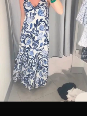 Blommig långklänning i blått och vitt - Säljer en långklänning med v-ringning och breda axelband. Klänningen har ett stort blommigt mönster i blått och vitt, samt en lätt utsvängd kjol. Perfekt för sommarens alla tillfällen!
