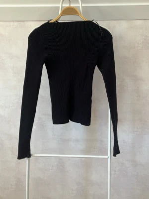 Svart ribbad stickad tröja - Säljer en svart, långärmad stickad tröja med ribbad struktur och snygga detaljer framtill. Tröjan har en kroppsnära passform och är tillverkad i ett stretchigt material som formar sig efter kroppen. Perfekt till jeans eller kjol.