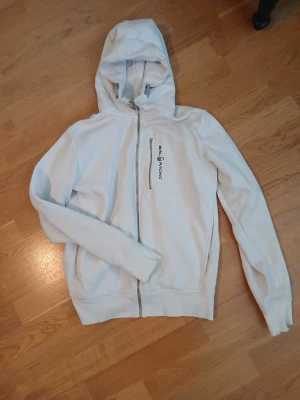 Ljusblå hoodie från Sail Racing - Ljusblå hoodie med dragkedja från Sail Racing. Tröjan har huva, ribbade muddar och en bröstficka med dragkedja. Märkets logga syns på bröstet. Perfekt för en avslappnad och sportig stil.