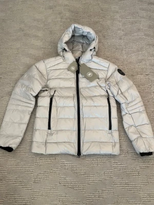 Silver pufferjacka från Canada Goose - Snygg silverfärgad pufferjacka från Canada Goose med huva och svarta detaljer. Jackan har två stora dragkedjeförsedda fickor framtill och logotyp på ärmen. Tillverkad i glansigt material och fylld för extra värme. Perfekt för våren 49 cm i axelbredd och 69 cm i längd