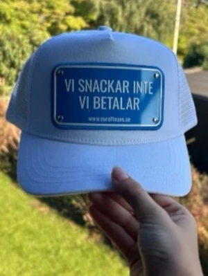 Vit keps från John Hatter - Cool vit keps från John Hatter med mesh på sidorna och bak. Framtill sitter en blå skylt med texten 'Vi snackar inte vi betalar'. Justerbar passform med snapback och tillverkad i polyester. Perfekt för dig som vill sticka ut med ett statement.