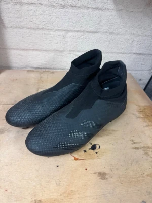 Svarta fotbollsskor Adidas Predator - Säljer ett par svarta Adidas Predator fotbollsskor med hög krage och mönstrad ovansida. Skorna har en strömlinjeformad design och diskreta svarta ränder på sidan. Materialet är syntetiskt och skon är utan snörning för en tight passform.