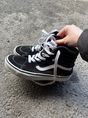 Svarta Vans Sk8-Hi sneakers - Säljer ett par klassiska Vans Sk8-Hi sneakers i svart med vita detaljer och vit sula. Skorna har högt skaft, vit sidostripe och vita snören. Materialet är canvas och mocka, perfekt för dig som gillar streetstyle och skate. Storlek 35 