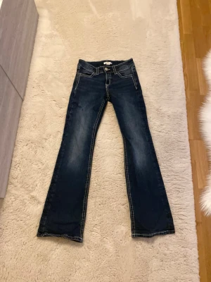  jeans från Ginatricot - Mörkblå bootcut jeans från Ginatricot med kontrastsömmar och klassisk femficksmodell. Jeansen har en något utsvängd passform nedtill och är tillverkade i denim med snygga slitningar framtill. Perfekta för en trendig och avslappnad look.