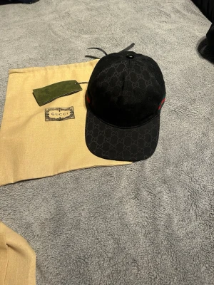 Svart Gucci keps med GG-mönster - Säljer en svart keps från Gucci med det klassiska GG-monogrammönstret över hela kepsen. På sidorna finns de ikoniska gröna och röda ränderna. Kepsen är i tygmaterial och har en böjd skärm. Perfekt för dig som vill ha en stilren och lyxig accessoar.